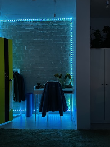 The image features a room with a modern aesthetic, highlighted by a brick wall illuminated with blue and green LED strip lights. A small desk is positioned against the wall with a chair draped in a coat. To the left, a bright yellow wardrobe contrasts against the dark, ambient lighting. A few plants and sunflowers in a vase are placed on the desk, adding a natural element to the decor.