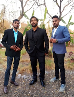 Three men dressed in formal attire are standing outdoors on a pebbled surface with dried grass and sparse plants visible. They are wearing blazers, two with black and one with a blue suit. The background shows trees and green banana plants, suggesting a natural setting.