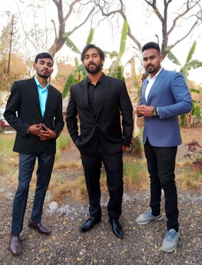 Three men dressed in formal attire are standing outdoors on a pebbled surface with dried grass and sparse plants visible. They are wearing blazers, two with black and one with a blue suit. The background shows trees and green banana plants, suggesting a natural setting.