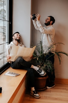 Two people are indoors, one is holding a camera and smiling as he looks to take a picture, while the other is sitting by a window with a map in hand, also smiling. A potted plant is positioned between them on the wooden floor. Light from the window illuminates part of the scene, and a magazine rests on the bench beside the seated person.