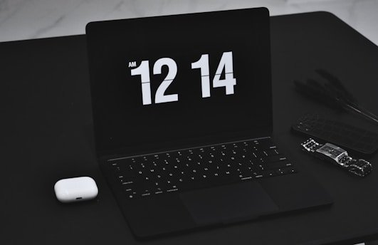 A sleek black laptop displays the time 12:14 AM in a bold, white digital font. Next to the laptop, on the left, is a pair of white wireless earbuds. On the right side, a smart watch and a cellphone with a dark case are positioned, adding to the tech-focused collection on a black table surface.