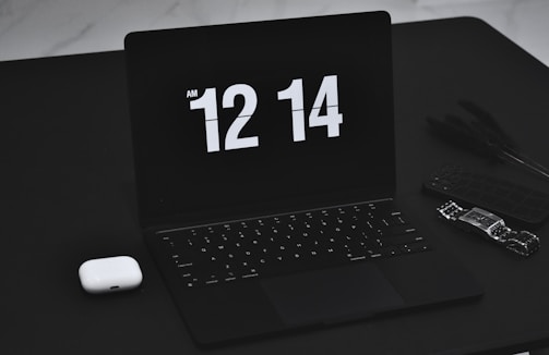 A sleek black laptop displays the time 12:14 AM in a bold, white digital font. Next to the laptop, on the left, is a pair of white wireless earbuds. On the right side, a smart watch and a cellphone with a dark case are positioned, adding to the tech-focused collection on a black table surface.