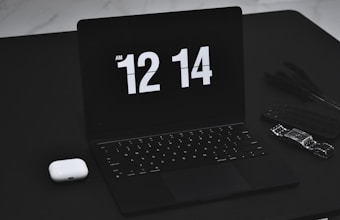 A sleek black laptop displays the time 12:14 AM in a bold, white digital font. Next to the laptop, on the left, is a pair of white wireless earbuds. On the right side, a smart watch and a cellphone with a dark case are positioned, adding to the tech-focused collection on a black table surface.