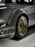 Close-up view of a sleek, modified sports car featuring prominent decals and logos such as 'Castrol' and 'Goodyear.' The image highlights the golden alloy wheel with an intricate mesh design, surrounded by a black tire. The car's body exhibits a metallic gray finish with riveted fender flares.
