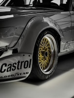 Close-up view of a sleek, modified sports car featuring prominent decals and logos such as 'Castrol' and 'Goodyear.' The image highlights the golden alloy wheel with an intricate mesh design, surrounded by a black tire. The car's body exhibits a metallic gray finish with riveted fender flares.