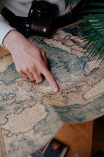 A hand points at a location on a detailed vintage map. A camera is partially visible, suggesting an interest in travel or exploration. Green leaves frame part of the image, adding a natural element.