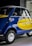 A small vintage car with a vibrant blue and yellow paint job, featuring the logo of a well-known airline company on its side. The car has a distinctive curved design with chrome accents, round headlights, and small tires. It is parked indoors, likely in a showroom or museum.