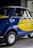 A small vintage car with a vibrant blue and yellow paint job, featuring the logo of a well-known airline company on its side. The car has a distinctive curved design with chrome accents, round headlights, and small tires. It is parked indoors, likely in a showroom or museum.