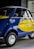 A small vintage car with a vibrant blue and yellow paint job, featuring the logo of a well-known airline company on its side. The car has a distinctive curved design with chrome accents, round headlights, and small tires. It is parked indoors, likely in a showroom or museum.
