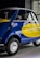 A small vintage car with a vibrant blue and yellow paint job, featuring the logo of a well-known airline company on its side. The car has a distinctive curved design with chrome accents, round headlights, and small tires. It is parked indoors, likely in a showroom or museum.