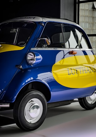 A small vintage car with a vibrant blue and yellow paint job, featuring the logo of a well-known airline company on its side. The car has a distinctive curved design with chrome accents, round headlights, and small tires. It is parked indoors, likely in a showroom or museum.