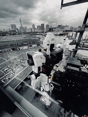 A large, white robot figure stands prominently on a platform near a waterfront, with a cityscape in the background. The sky is overcast, casting a gray tone over the scene. The robot, resembling a giant mecha, has intricate details and appears to be a popular attraction with people gathered at its base.