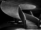 A dramatic spiral staircase crafted from concrete, showcasing its curves and lines against a stark, minimalist background. The structure elegantly rises with its smooth, sweeping design, creating a sense of movement and flow. Shadows and light interplay to highlight the architectural details.