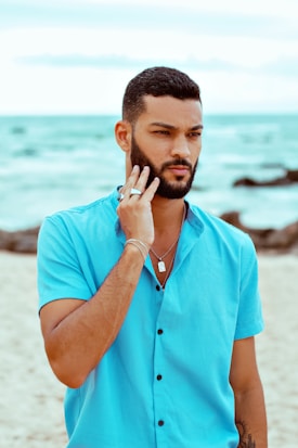 A man with short hair and a beard is standing on a beach. He is wearing a bright turquoise short-sleeved shirt and silver jewelry, including a necklace and multiple rings. The background features a cloudy sky, waves in the sea, and some rocks.