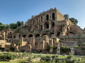 An ancient Roman ruin with multiple arches and weathered stone walls set amid lush green foliage. The foreground features a garden with statues and rose bushes, while people can be seen walking along the pathways attached to the structure. The scene is bathed in warm sunlight under a clear blue sky.