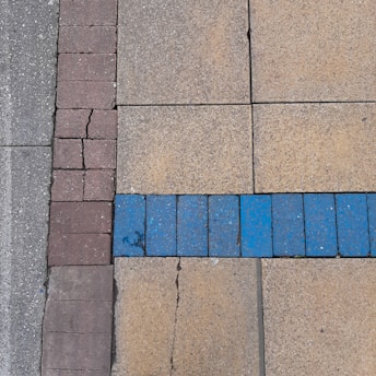 A section of pavement with variously colored tiles. The surface is divided into three parts: gray concrete on the far left, a panel of small reddish-brown tiles adjacent to large beige tiles, with an uneven line of blue tiles crossing horizontally. The tiles appear slightly worn and cracked.
