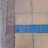 A section of pavement with variously colored tiles. The surface is divided into three parts: gray concrete on the far left, a panel of small reddish-brown tiles adjacent to large beige tiles, with an uneven line of blue tiles crossing horizontally. The tiles appear slightly worn and cracked.