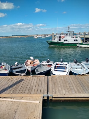 A row of small boats is moored along a wooden dock. The boats have outboard motors and are positioned on calm water under a blue sky with scattered clouds. In the background, a larger green and white vessel is on the water, with a pier and an expansive view of the sea and distant horizon.