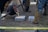 Molten metal is being poured from a crucible into a mold situated on the ground. Several individuals in protective clothing, including heavy-duty boots, leggings, and aprons, are overseeing and facilitating the process. The scene appears to be outdoors, with a visible patch of grass and dirt.