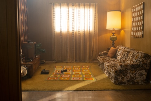 A retro-style living room is depicted with dim lighting. The wooden-paneled walls give the room a warm atmosphere. A vintage floral-patterned couch is placed against the right wall, next to a tall, ornate lamp. The floor is covered with a yellow shag carpet with a square-printed area rug in the center featuring yellow and red patterns. The window is covered with sheer curtains, and there is a wooden console with a TV on the left side of the room. Another piece of art with geometric patterns hangs on the right wall.