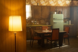 A vintage kitchen scene featuring wooden cabinets and a retro-style mint green refrigerator. The room is adorned with floral wallpaper in warm tones. A wooden dining table with four chairs is positioned in the center, accompanied by a set table with tableware. A tall floor lamp with a white shade emits a warm glow, casting a cozy ambiance across the room.