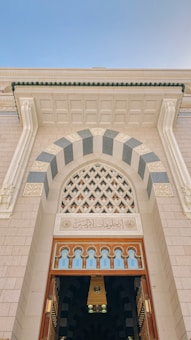 An ornate building facade with a large arched entrance, featuring intricate geometric patterns and a blend of light and dark stone. Above the wooden doors, there is an inscription in Arabic script.