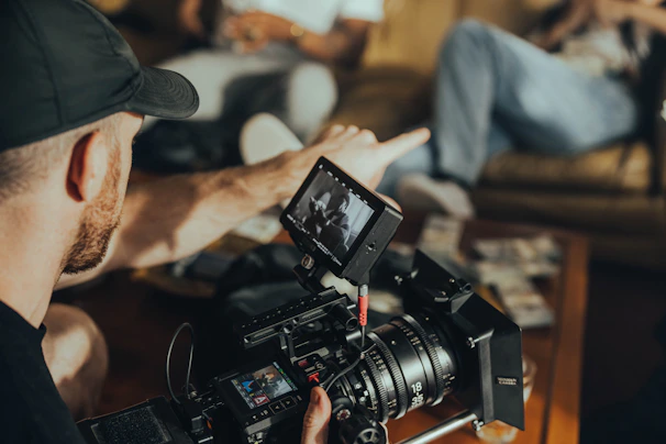 A person is operating a professional video camera indoors. The focus is on the camera and its mounted monitor displaying a scene. Another individual is sitting on a couch in the background, partially out of focus.