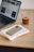 A laptop displaying a webpage is placed on a wooden desk alongside a glass of coffee. Nearby, a smartphone with a case rests on top of a couple of notebooks with illustrations.