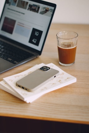 A laptop displaying a webpage is placed on a wooden desk alongside a glass of coffee. Nearby, a smartphone with a case rests on top of a couple of notebooks with illustrations.