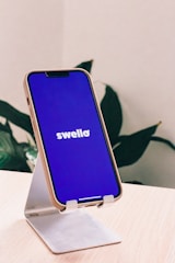 A smartphone with a blue screen displaying the word 'swello' rests on a white stand. The background features green leaves, indicating a plant in an indoor setting. The phone case appears to be tan, and it is placed on a light wooden surface.