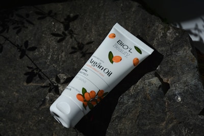 A tube of Bio'l Beauty Argan Oil cream lies on a dark rock surface, surrounded by shadows of leaves. The packaging features orange fruit illustrations, suggesting a natural theme.