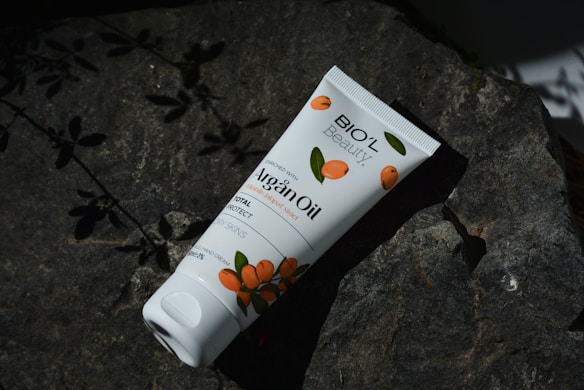 A tube of Bio'l Beauty Argan Oil cream lies on a dark rock surface, surrounded by shadows of leaves. The packaging features orange fruit illustrations, suggesting a natural theme.