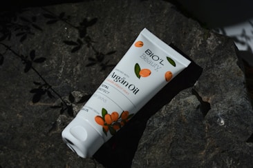 A tube of Bio'l Beauty Argan Oil cream lies on a dark rock surface, surrounded by shadows of leaves. The packaging features orange fruit illustrations, suggesting a natural theme.
