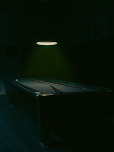 A dimly lit pool table rests in a dark room with only a single overhead light illuminating it. A cue ball and a pool cue are positioned on the table, casting soft shadows on the felt surface. The atmosphere is quiet and still, with a focus on the pool table as the central object.