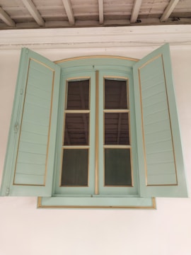 A vintage-style window with shutters painted in a soft green color. The window features clear glass panes divided by wooden muntins, set within a light wooden frame that exhibits subtle golden detailing. The shutters are open, revealing the contrasting dark interior. Above the window, the ceiling has exposed wooden beams, painted in an off-white or light gray shade.