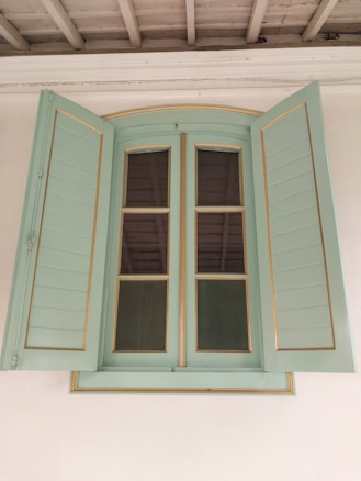 A vintage-style window with shutters painted in a soft green color. The window features clear glass panes divided by wooden muntins, set within a light wooden frame that exhibits subtle golden detailing. The shutters are open, revealing the contrasting dark interior. Above the window, the ceiling has exposed wooden beams, painted in an off-white or light gray shade.
