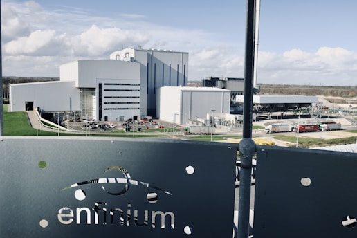 A large industrial facility with multiple modern, white and grey buildings surrounded by roads and green lawns. The foreground features a metallic fence with the word 'enfinium' visible. Trucks and other vehicles are near the buildings.