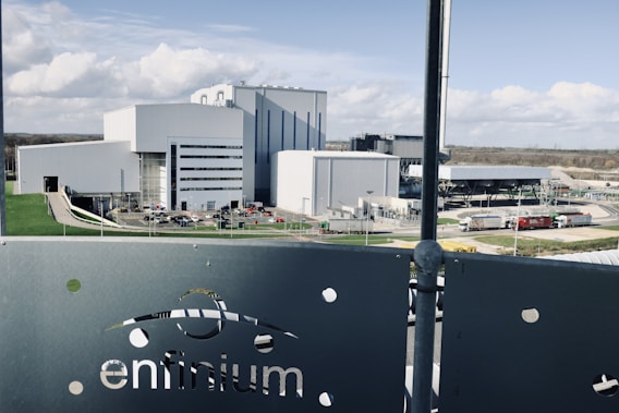 A large industrial facility with multiple modern, white and grey buildings surrounded by roads and green lawns. The foreground features a metallic fence with the word 'enfinium' visible. Trucks and other vehicles are near the buildings.