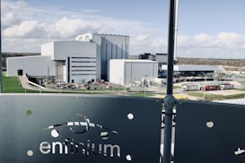 A large industrial facility with multiple modern, white and grey buildings surrounded by roads and green lawns. The foreground features a metallic fence with the word 'enfinium' visible. Trucks and other vehicles are near the buildings.