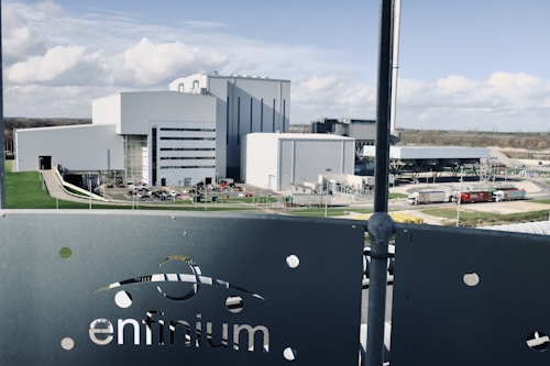 A large industrial facility with multiple modern, white and grey buildings surrounded by roads and green lawns. The foreground features a metallic fence with the word 'enfinium' visible. Trucks and other vehicles are near the buildings.