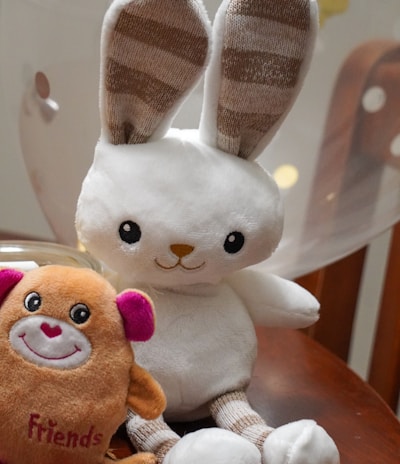A plush toy rabbit with long ears and striped inner ear patterns sits on a wooden surface next to a small plush toy in the shape of a creature with pink ears and the word 'Friends' embroidered on it. A balloon softly blurred in the background adds to the playful setting.
