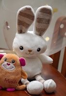 A plush toy rabbit with long ears and striped inner ear patterns sits on a wooden surface next to a small plush toy in the shape of a creature with pink ears and the word 'Friends' embroidered on it. A balloon softly blurred in the background adds to the playful setting.