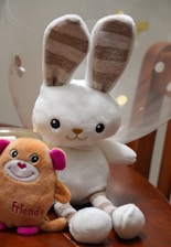 A plush toy rabbit with long ears and striped inner ear patterns sits on a wooden surface next to a small plush toy in the shape of a creature with pink ears and the word 'Friends' embroidered on it. A balloon softly blurred in the background adds to the playful setting.