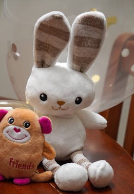 A plush toy rabbit with long ears and striped inner ear patterns sits on a wooden surface next to a small plush toy in the shape of a creature with pink ears and the word 'Friends' embroidered on it. A balloon softly blurred in the background adds to the playful setting.