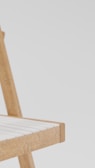 A close-up view of the corner of a wooden chair with a light-colored seating surface. The chair has a simple and minimalist design with smooth, straight lines and natural wood texture. The background is plain and light-colored, providing contrast and focus to the chair.
