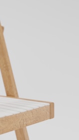 A close-up view of the corner of a wooden chair with a light-colored seating surface. The chair has a simple and minimalist design with smooth, straight lines and natural wood texture. The background is plain and light-colored, providing contrast and focus to the chair.