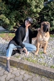 A person dressed in casual clothing, including a black cap and jeans, is sitting on the edge of a curb next to a large, brown dog. The person has one arm resting on the dog and is holding a small item in the other hand. They are outdoors, with a background of greenery, mulch, and rocks.