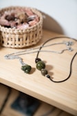 A wooden surface holds a necklace with large green and brown beads, alongside a silver chain. A wicker basket filled with various small items, possibly seashells or dried flowers, is in the background.