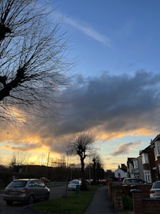 A suburban street at sunset with bare trees lining the sidewalk. A few parked cars are visible on the left, and houses with brick facades are on the right. The sky is filled with dramatic clouds, illuminated by the golden light of the setting sun.