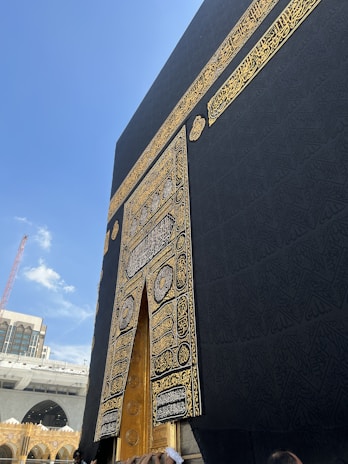 A close-up view of the Kaaba's door, adorned with intricate gold calligraphy and patterns on a black cloth. The Kaaba, located in the Grand Mosque of Mecca, is surrounded by a clear blue sky and nearby architectural structures. Several people are visible near the bottom of the image, seemingly in an act of worship or reverence.